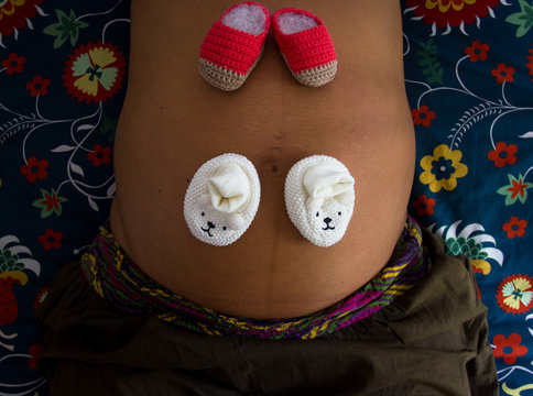 Closeup Baby Knitted Shoes On Naked Big Belly Of Pregnant Woman. Mother Lying On Colorful Duvet Cover Bed. Twins, Siblings, Newborn Concept