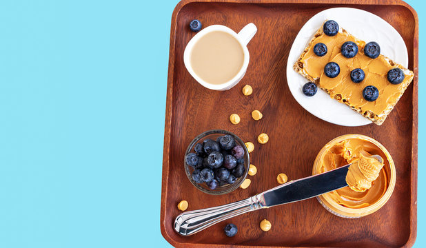 Peanut butter concept healthy breakfast. Sandwich with multigrain crisp with peanut butter and blueberries and cup of cappuccino coffee. Top view. Wooden plate blue pastel backround.