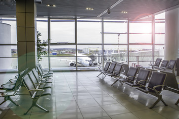 Airplane, view from airport terminal. Modern airport terminal with black leather seats on a sunny morning. A huge viewing glass facade with a passenger aircraft behind it.