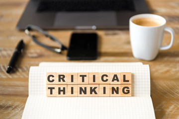 Closeup on notebook over wood table background, focus on wooden blocks with letters making Critical Thinking text