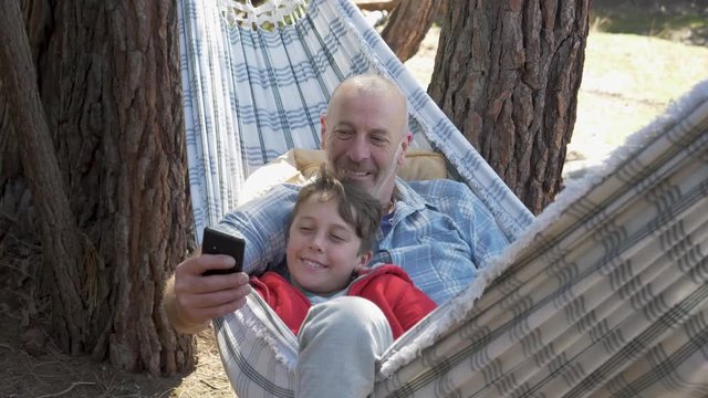 Father and son taking cell phone selfies together in a hammock in nature