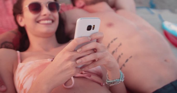 Hipster couple sunbathing at the beach and texting on mobile-phone