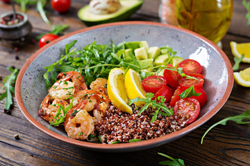 Delicious healthy Buddha bowl with shrimps, tomato, avocado, quinoa, lemon and arugula on the wooden table. Healthy food.
