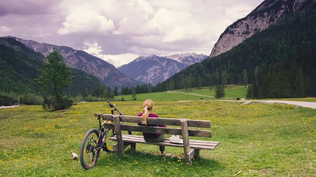 Girl sits on bench in front of beautiful landscape from forest, field and mountains. Rest during bike trip in the Alps. Meditation in heavenly place. Cycling in the mountains.