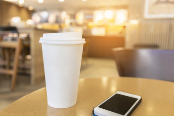 Disposable hot coffee cup with smartphone on a wooden table in coffee shop background