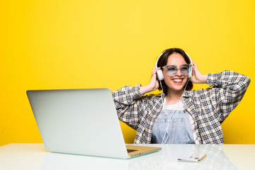 Cheerful young woman in headphones dancing to music while sitting in front of computer on yellow background