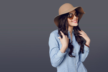 A beautiful brunette smiling young woman with curly long hair posing in hat and sunglasess, isolated on grey background. Horizontal view.