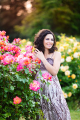 Obraz na płótnie Canvas A vertical portrait of young Caucasian woman with dark brown curly hair near pink rose bushes, looking to her left, smiling with teeth, in summer dress