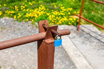 Blue lock on old rusty car ramp, with blurred yellow dandelions on spring meadow in background.