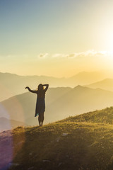 girl in dress with hands up standing on grass in sunset mountains
