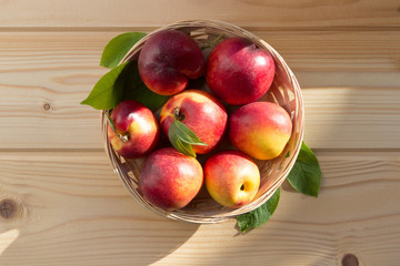 fresh nectarine with leaves  are in a wicker plate on a light wooden table in the sun. close-up