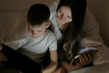 Young woman mom and her son watching interesting film together on tablet and laugh under the blanket. Mom with phone.