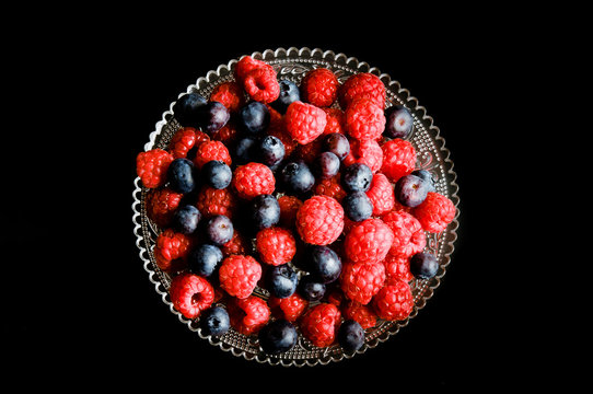 red raspberries and bluberries berries on glass plate over black background 