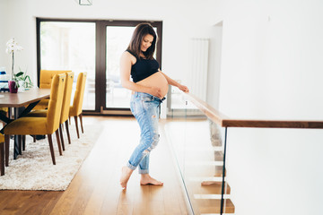Beautiful pregnant woman relaxing, touching and smoothing her tummy, smiling. New home, interior design and living room
