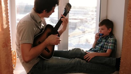 Playing a musical instrument. Dad is playing the guitar and son is playing mouth organ sitting in windowsill.