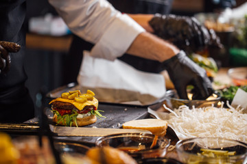 Process of cooking burgers. Cropeed view of chef hands in black gloves with prepared cheeseburger, variety of fillings and ingridients on wooden desk. Catering and Good Quality Fastfood concept