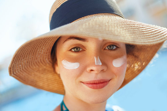 Portrait of a beautiful young woman, smiling, in a swimsuit, smeared face with sun protection cream, hat, sea background. Female Applying Sun Cream on face.