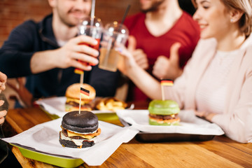 leisure, celebration, drinks and holidays concept - happy people and friends clinking glasses of beverage at burger bar. Different burgers served on wooden table.
