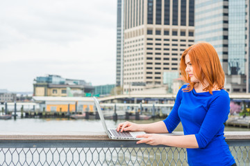 Young American Woman traveling, working in New York, wearing blue long sleeve T shirt, standing by river at harbor in business district of Manhattan, looking down, working on laptop computer..