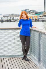 Young American Woman traveling in Manhattan, New York, wearing blue long sleeve T shirt, long black leather boots, standing by river at harbor under cloudy sky, looking forward, relaxing, thinking. .