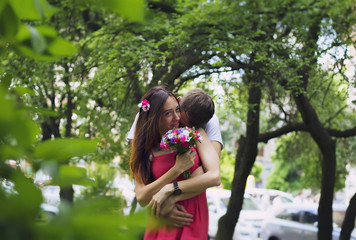 Couple in love are hugging and kissing in the green summer park. Girl in pink dress is holding rustic bright bouquet in her hands. Trees and nature outdoors.