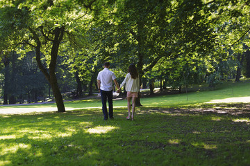 Couple in love is walking away holding hands in the green summer park. Trees and nature outdoors. Girl in short pink dress and guy in smart suit.