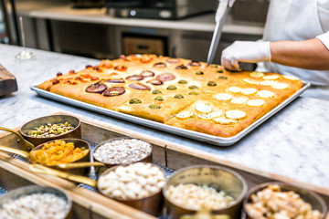 Restaurant kitchen cutting foccacia bread with knife, nuts, spices in containers and toppings man chef after baking in oven italian