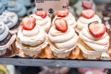 Macro closeup of vanilla strawberry cupcakes on display in bakery in muffin paper liners with white cream cheese frosting icing, cut fresh berries