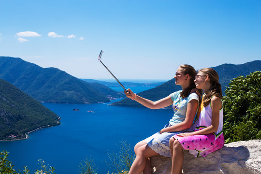 Two teenage girls in the make photo on the phone and selfie sticks on the background of Boko Kotorska bay and mountains in Montenegro.