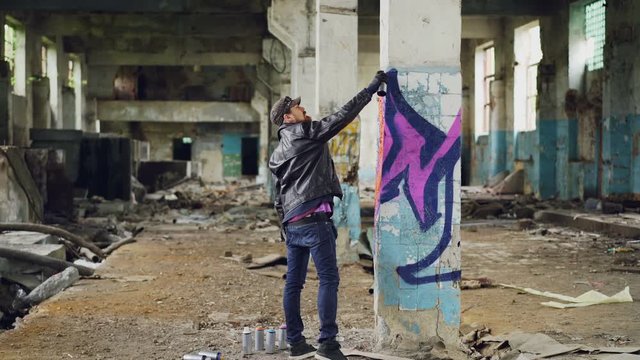 Graffiti painter in casual clothing is drawing with spray paint on column in spacious abandoned building. Abstract images, modern art, creative people and hipsters concept.