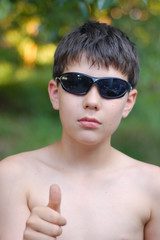 Fototapeta premium Young boy in the glasses from the sun resting in nature and showing the class with a thumb of his hand. He loves to relax with his parents in the nature