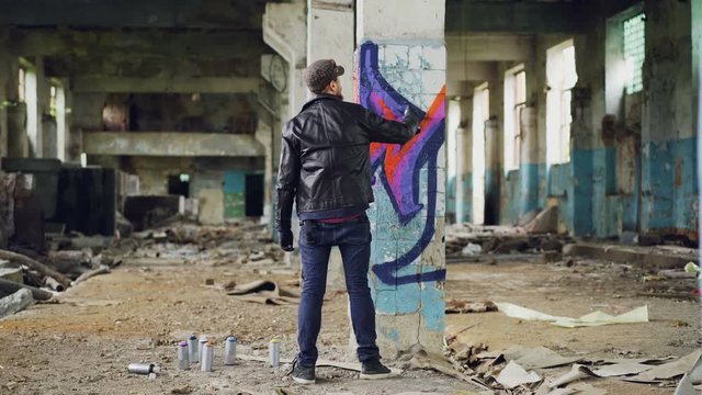 Back view of graffiti painter creating beautiful image with aerosol paint inside abandoned building. Artist is wearing blue jeans, black leather jacket, cap and gloves.