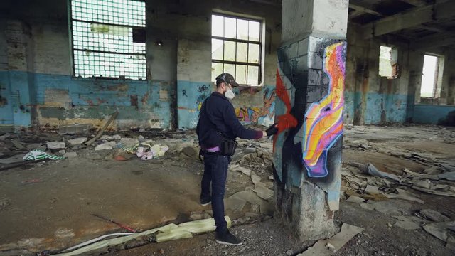 Male graffiti artist is decorating old damaged column inside empty industrial building with abstract pictures. Modern painter is using aerosol spray paint.