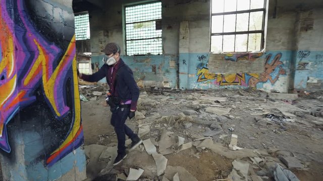 Urban street artist is decorating old pillar inside abandoned house with dirty walls and windows, he is using paint spray. Modern artwork and creative people concept.
