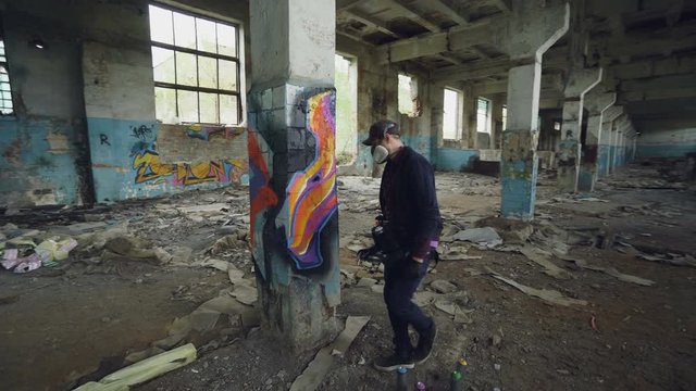 Graffiti painter is protective mask and gloves is drawing on old column in dirty empty building using aerosol paint. Young man is wearing casual clothes and cap.