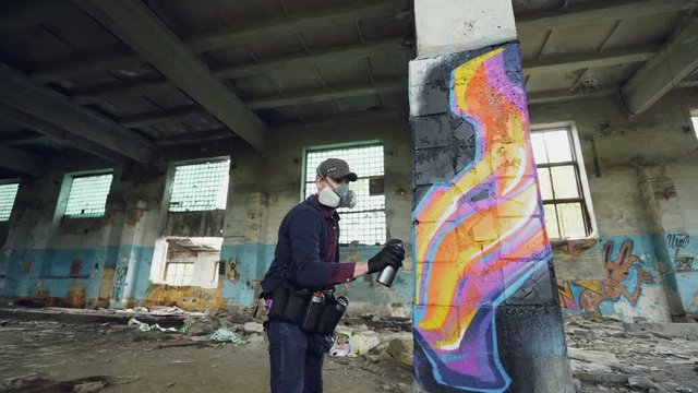 Low angle shot of young man is face mask painting graffiti on column inside empty industrial building using spray paint. Dirty damaged walls, windows and ceiling are visible.