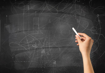 Fototapeta premium Female hand holding white chalk in front of a blackboard with scribbles and plans drawn on it 