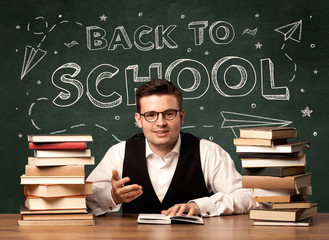 A young teacher in glasses sitting at classroom desk with pile of books in front of blackboard saying back to school drawing concept.