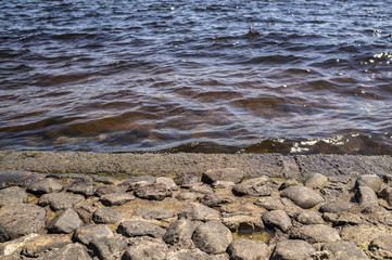 An ancient embankment of cobblestone in the background of a river with clear water and waves in the rays of the spring sun.