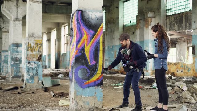 Experienced graffiti artist is teaching his friend to create beautiful images with spray paint, they are standing together in adandoned building near old column and talking.