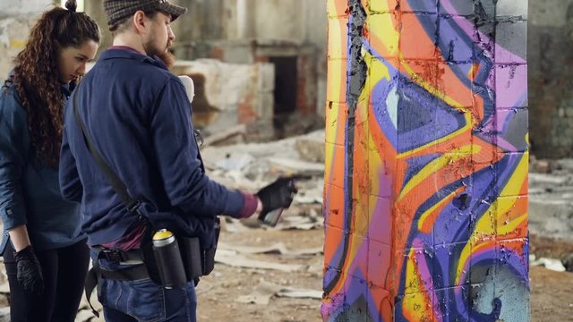 Urban artist handsome bearded man is teaching amateur student to work with aerosol paint while decorating old industrial building with abstract graffiti.