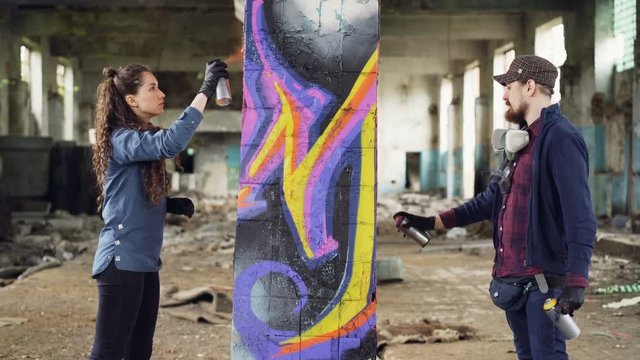 Two skilled graffiti artists bearded guy and attractive young woman are working together in abandoned warehouse decorating old column with abstract image.