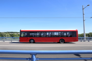 City bus crossing bridge. Red bus moving on road against blue barrier or guard rail.