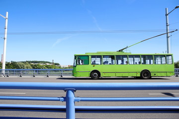 Fototapeta premium Trolley bus crossing bridge. Green trolleybus moving on road against blue barrier or guard rail.
