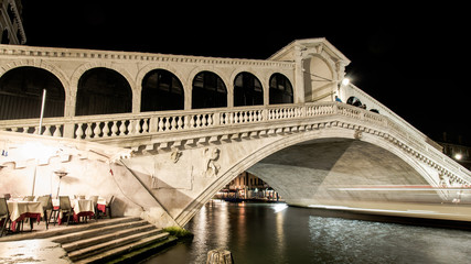 Ponte Rialto, the Royal Bridge, spanning the Grand Canal in Venice at night. Streaks of light pass under the bridge from a passing water taxi