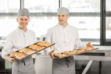 Portrait of a two bakers standing with tray full of freshly baked buns at the manufacturing