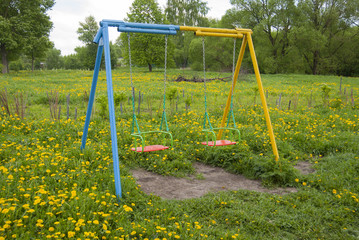 Swings for children, empty, yellow-blue, against a background of bright green grass with yellow dandelions