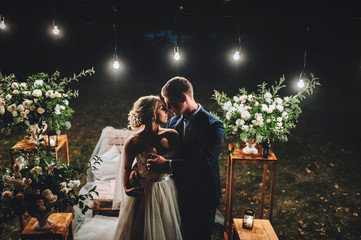 Fashionable newlyweds stand at night against the backdrop of chic decorations and flowers, electric bulbs and garlands, in the backlight. Night shooting of the  bride and groom. Floristic. Film effect
