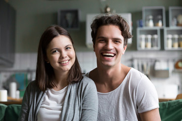 Happy millennial couple looking at camera in the kitchen, smiling attractive young man and woman posing at home, cheerful casual teenage boyfriend and girlfriend sitting on couch headshot portrait