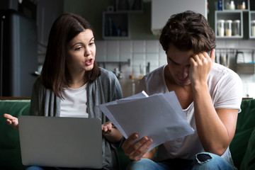 Angry wife scolding blaming upset husband of unpaid bills, bankruptcy or past due debt, unhappy millennial couple having financial problems arguing at home sitting on sofa with laptop and papers